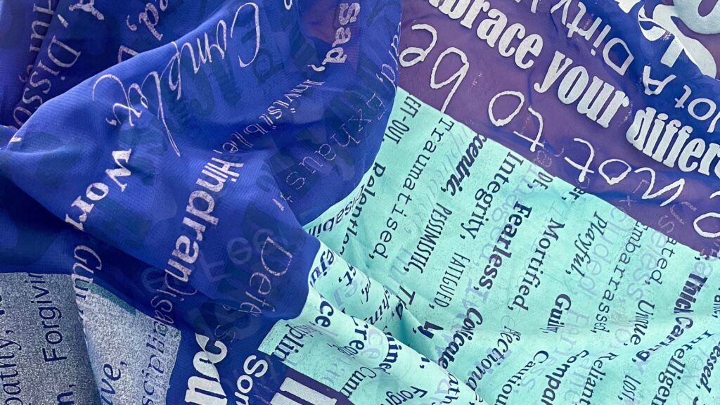 A close up photo of a pile of blue fabrics which have been printed onto. The fabrics are covered in a mixture of positive and negative words related to disability.