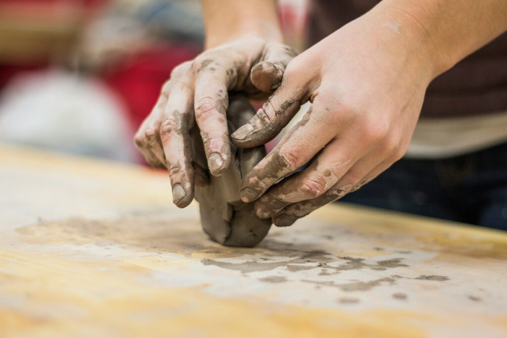 Hands shaping some clay with messy fingers and clay on a wooden table below.