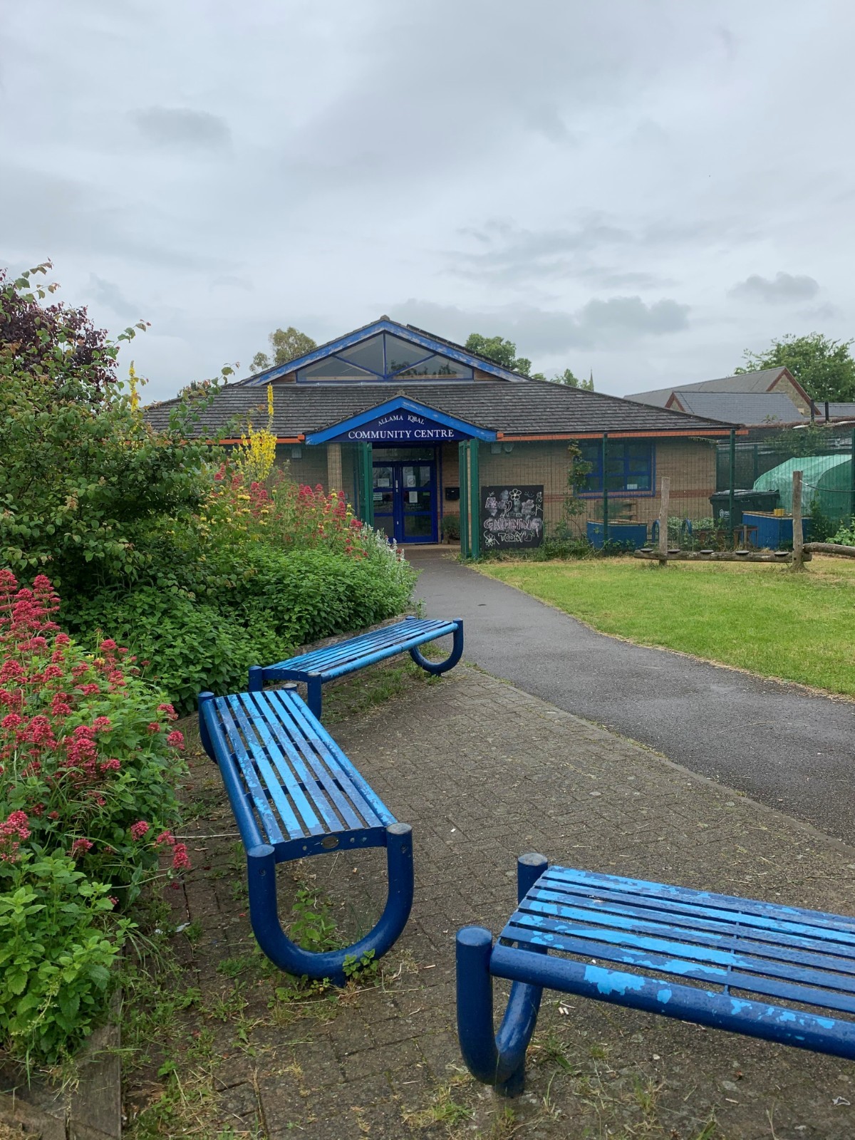 A photo of the front of the Allama Iqbal Centre. On the left is lush bushes in front of 3 blue benches, which is known as the Iqbal Pocket Park. The centre is at the end of the path and is a ground-floor community centre with a unique shaped roof.