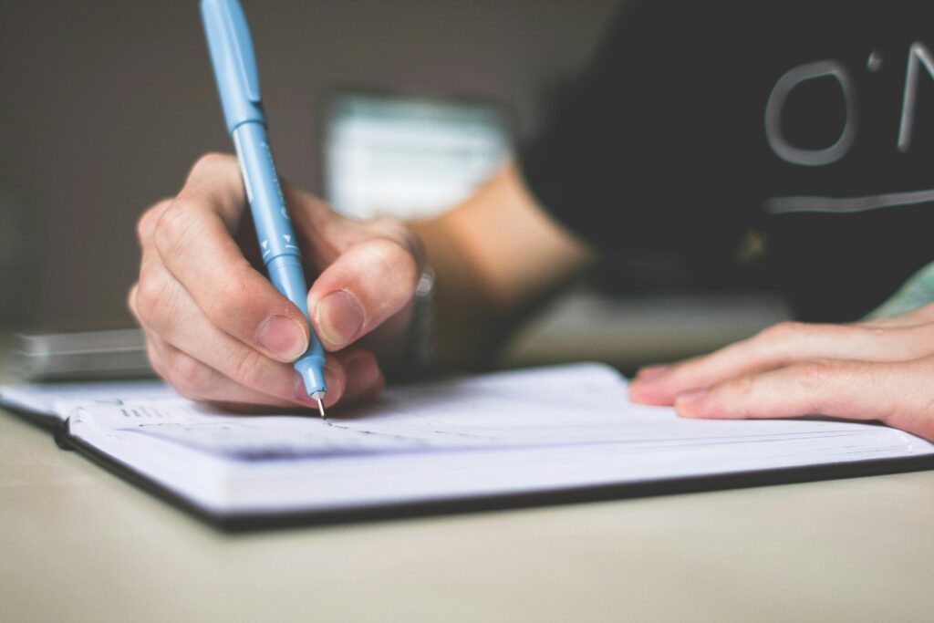 Close crop of hand holding a pale blue pen to a notebook placed on a table.