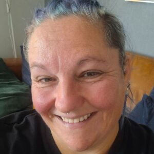 A portrait photo of Jen. Jen has brightly coloured hair tied back. She has a friendly, welcoming smile whilst looking at the camera. 