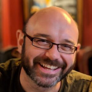 A portrait photo of Alan. Alan is wearing thick framed black glasses, has a beard and moustache and a friendly, warm smile.