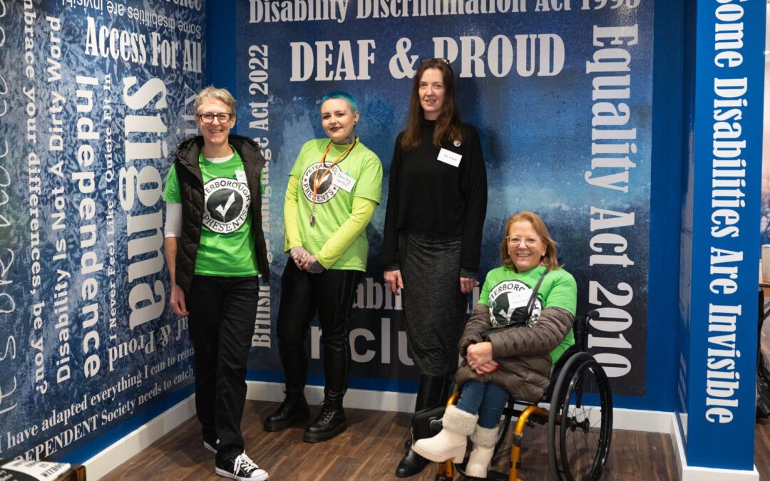 Four people posed in front of an exhibition wall with phrases and slogans around disability. The artist Fae is surrounded by three people wearing peterborough presents branded green tshirts.