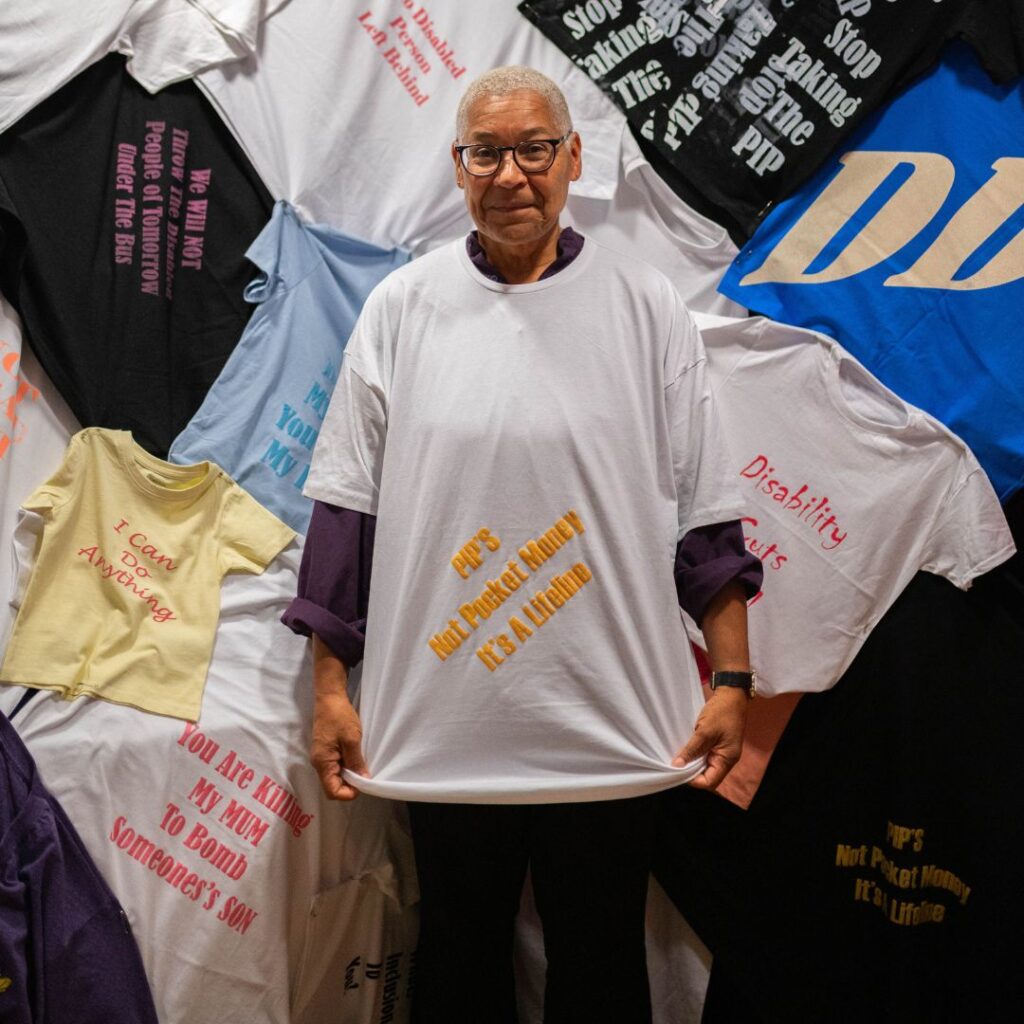 A person with short cropped hair and dark framed glasses stands in front of a wall of slogan T-shirts, smiling and wearing their own slogan shirt.