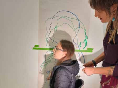 An example of what went on at the Art LAB. David, a teenager, sits with his profile parallel to a wall. His shadow cast on the wall is being drawn around by artist Rose. David has long brown hair, glasses and is wearing a padded, hooded navy coat
