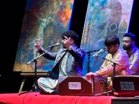 A close up photo of 3 of the members of Chand Ali Khan Qawwal and party performing.  They are seated cross legged on a red carpet, microphones pointing at their faces, dressed in shalwar kameez. The lead singer, Chand, has his arms raised as he is singing.