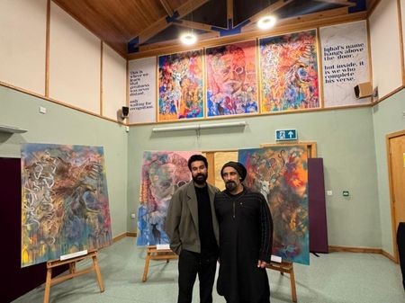Zain (left) and Seyyed (right) stand with their artwork collection on display. Directly behind them are large canvases displayed on easels, with colourful paintings of an eagle, a portrait of allama iwbal, and a white deer. The artworks have combined text and visual art, and have lines of urdu and english poetry woven between the illustrations. Above them, these artworks are repeated as vinyls attached to the wooden panels, alongside two verses of poetry on either side.