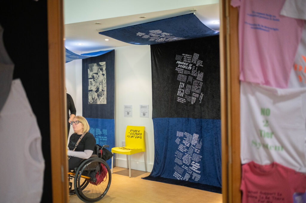 The Power of Protest and Print exhibition. A collection of fabric artworks predominantly in blues, hang from the ceiling and trail along the wall. There is unknown text printed on the large fabric panels. On the left, a blonde woman in a wheelchair is looking off to the left, appearing in conversation with someone out of frame.