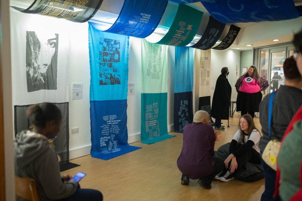 The Power of Protest and Print exhibition. A collection of fabric artworks predominantly in blues, hang from the ceiling and trail along the wall. There is unknown text printed on the large fabric panels. On the right on the image, a group of people are in conversation.