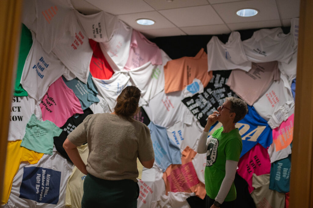 Two people stand in the Hundred Years of Protest Room, looking up at the t-shirts which are hanging from the ceiling and along the walls.