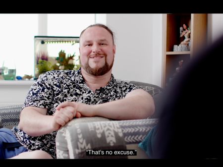 Sandy Wardrop screen grab from Jay's film. Sandy is resting his arm across the arm rest of a pale grey sofa, clasping his hands together. He is smiling while speaking, and has short brown hair and a short brown beard and mustache. The caption reads "That's no excuse".