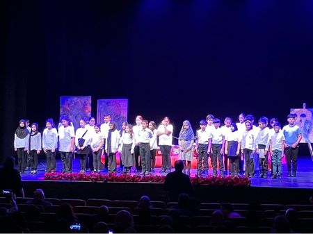 A photo of a large group of school children stood on a theatre stage. The children are all dressed in white shirts and dark trousers.