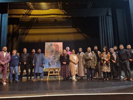 A large group of 20-30 people stood on stage at the new theatre. A large proportion of people in the picture are British Pakistani / South Asian disapora. In the middle of the group is a large canvas on an easel. The art is a colourful portrait of allama iqbal.