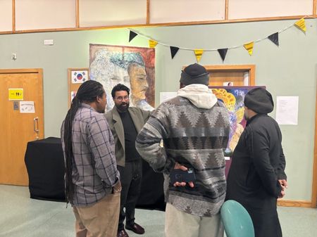 Zain and 3 audience members in conversation at the exhibition. Zain is looking at the group and pointing at the artwork, which is hidden from view behind the group.