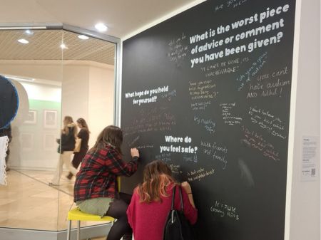 Two people writing on a chalkboard wall in the RAD Collections Exhibition. On the wall are lots of sentences written in colourful chalk, answering quetions about where people feel safe, and what's the worst piece of advice they've received.