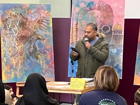 An asian man reading a poem at the open mic. He holds his phone in one hand and a mic in the other. Behind him, colourful paintings of allama iqbal and an eagle