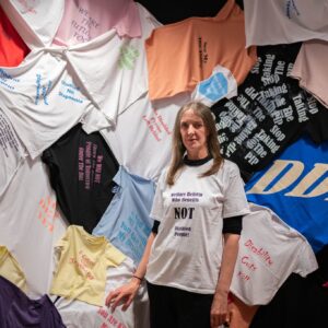 Artist Fae Kilburn stands in her "Hundred Years of Protest" t-shirt room. The wall is completely covered with an assortment of disability themed protest t-shirts. Fae is a tall woman with long dark hair. She is wearing a white t-shirt with protest slogan in large black font which says: Welfare Reform. Who Benefits? NOT Disabled People.