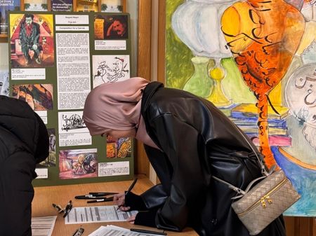 A photo of a young woman in a dusky pink silk hijab and black coat, leaning over as she fills in a form. Behind her, vibrant artwork with urdu script and curved shapes in orange, yellow, and green.