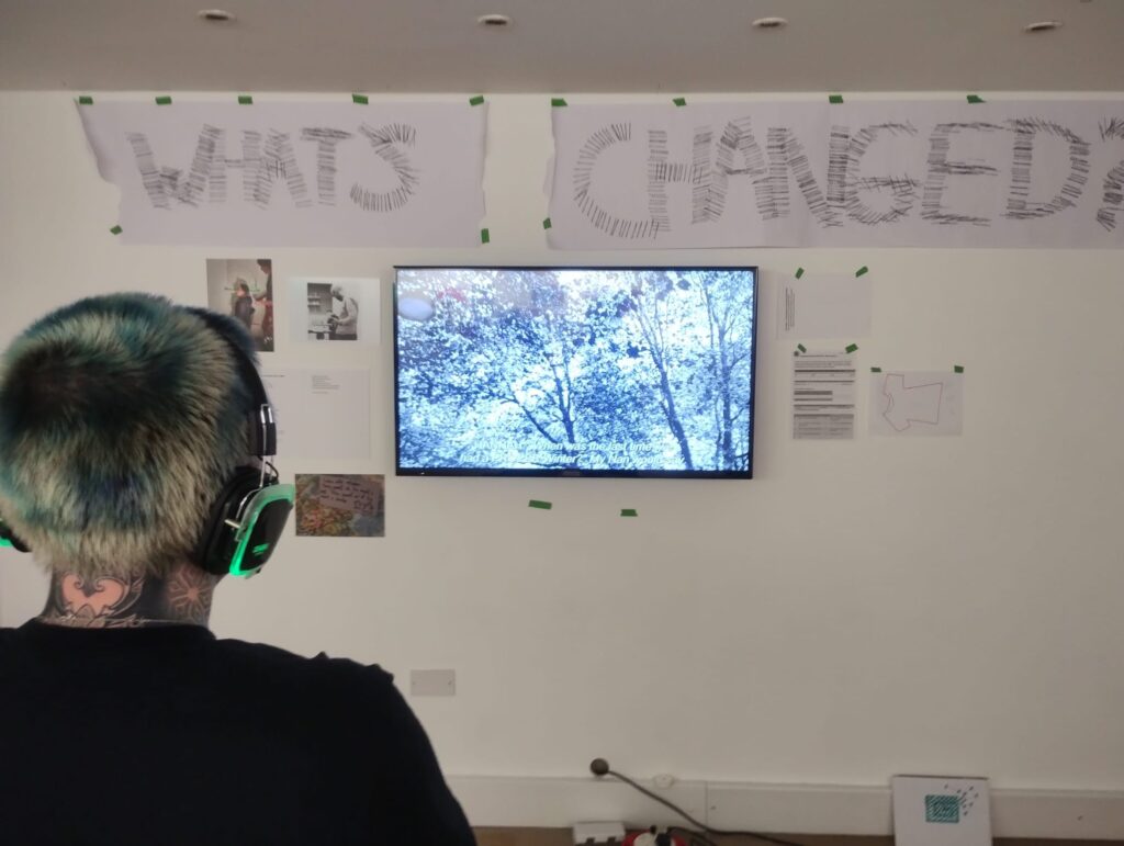 John's film in the RAD Collections exhibition. The wall has various papers stuck to it, around the tv mounted to the screen. Charley is looking at the screen and watching it, wearing wireless headphones