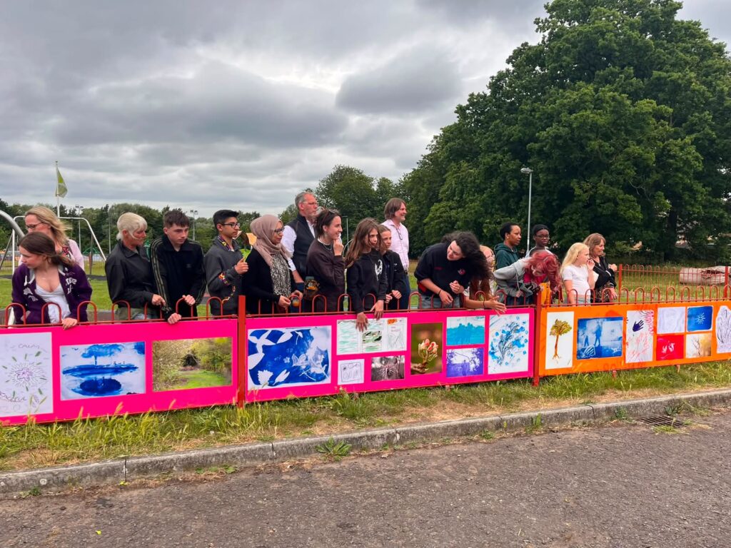 (approx 18) Eye villagers and the young people of Eye gathering for a photo with their Outdoor Exhibition that is colourfully on the railings in front of them.