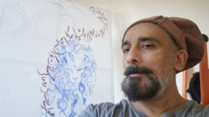 A selfie of Seyyed Naqvi in front of one of his works in progress. 
Seyyed is a brown man with a black and silver cropped bears and dark brown eyes. He is wearing a medium brown hat and a grey t-shirt. The artwork behind him is a work in progress coloured pencil sketch. The centre of the work is of a stylised illustration of a lion, with arabic calligraphy flowing round the lion in a dark brown colour.
