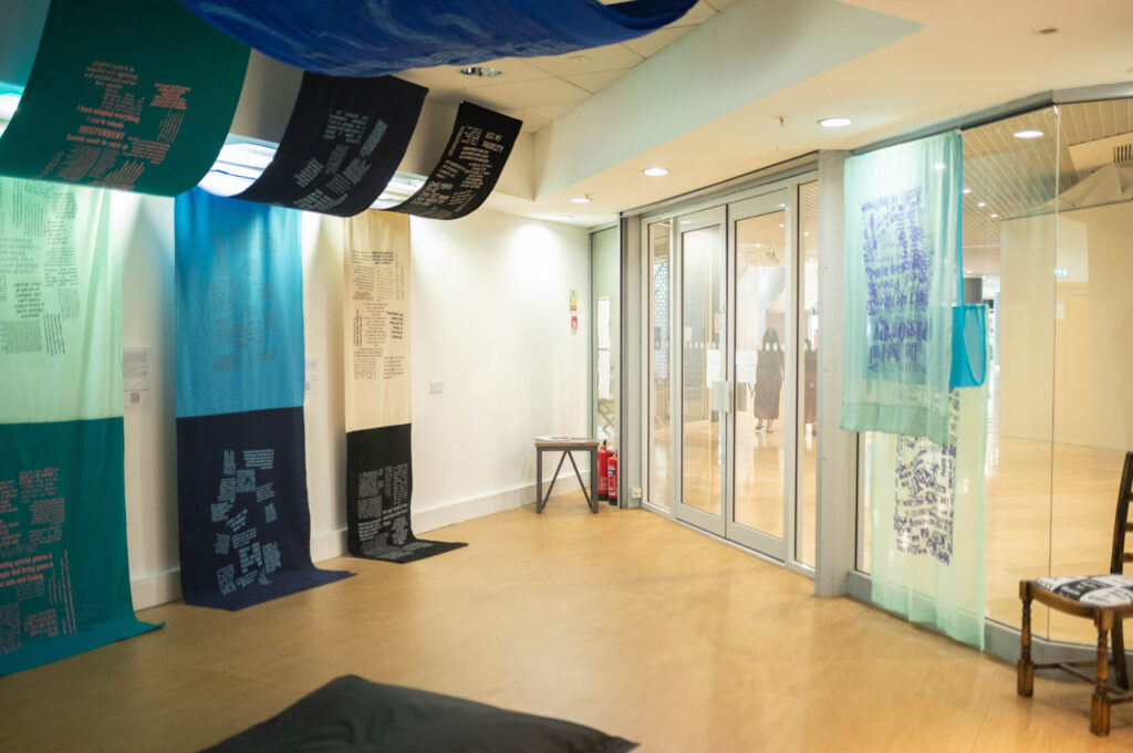 The Power of Protest and Print exhibition. A collection of fabric artworks predominantly in blues, hang from the ceiling and trail along the wall. There is unknown text printed on the large fabric panels.