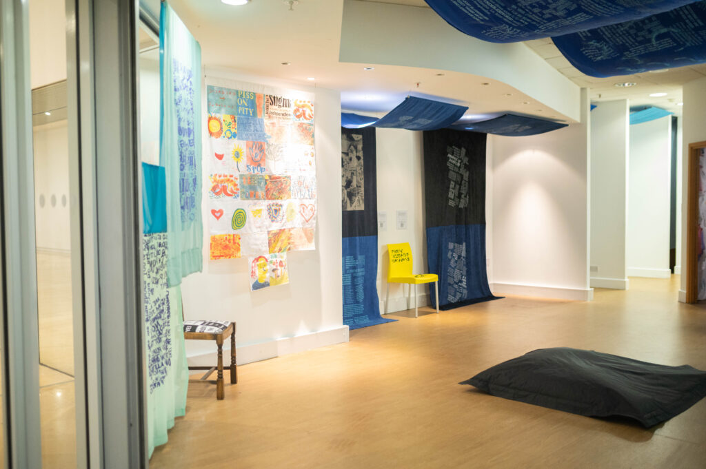The Power of Protest and Print exhibition. A collection of fabric artworks predominantly in blues, hang from the ceiling and trail along the wall. There is unknown text printed on the large fabric panels. A bright yellow shair separates the two blue panels furthest away.