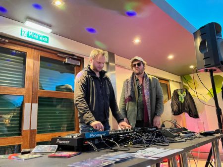 Two people stand at the DJ decks. The person on the left is spinning the decks and the person on the right is wearing sunglasses.