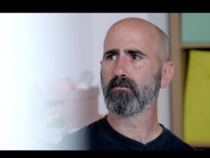 Jay looks to the left of the frame and has a calm, listening look on his face. He is a white man with a black and grey cropped beard, wearing a black t-shirt