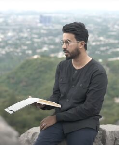Zain sits on a rock overlooking a vast landscape of pale greens and dusty grey buildings. Zain is a brown man with black hair and a short black beard and black, circle rimmed glasses. He is wearing a dark grey long sleeved jumper, and holding a book in his right hand. He is looking to the left as he recites his poetry.