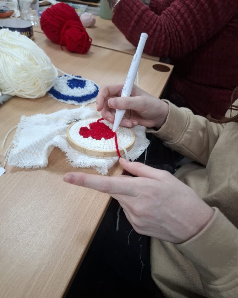 Close up of someone making a needle punch design on a craft hoop with yarn. The design is a red heart on a white background. The person crafting is in the process, with one hand on the needle punch and the other holding the hoop and yarn. 