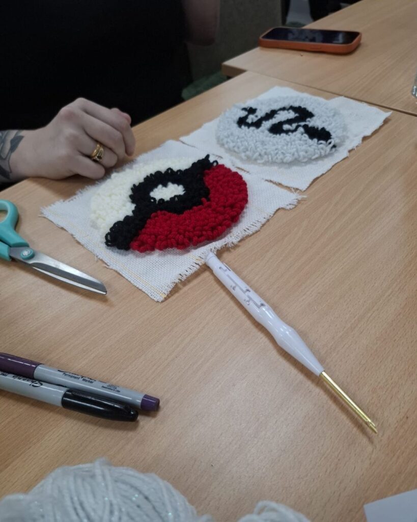 A close up of two needle punch designs laid on a wooden table. Both designs are circular, with one looking like a pokeball and the other a black and white snake design. There are craft supplies scattered on the table.