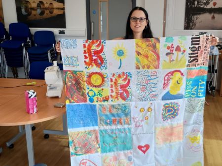 Dee, a woman with long black hair and thick black rimmed glasses, holding up the patchwork community panel art. To her right on the table is a sewing machine, which suggests she has just sewn this colourful patchwork panel together.