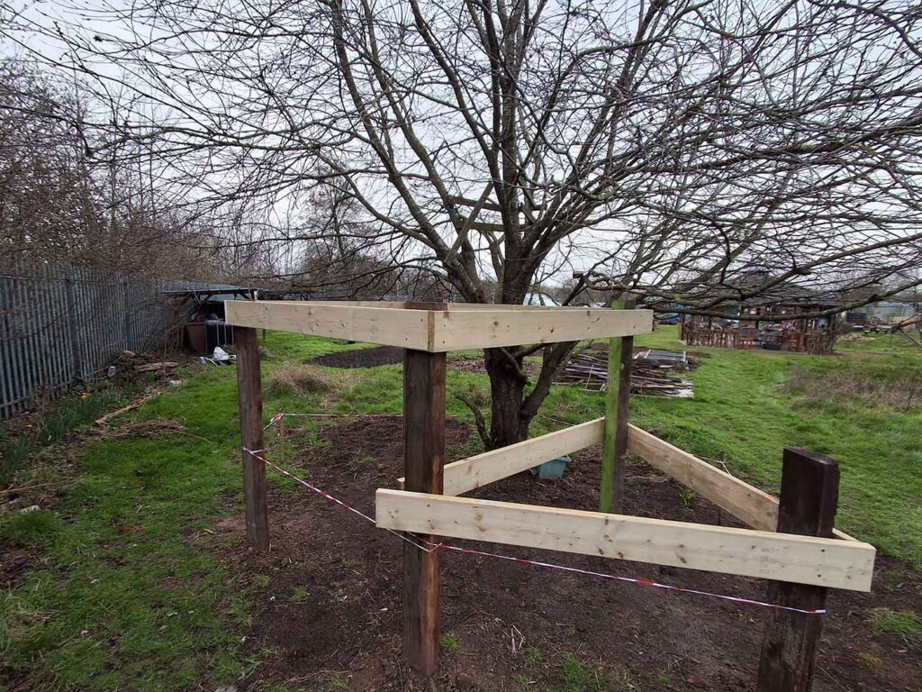 A wooden structure with two levels, the start of a building, freshly built in a green outdoor space with trees.