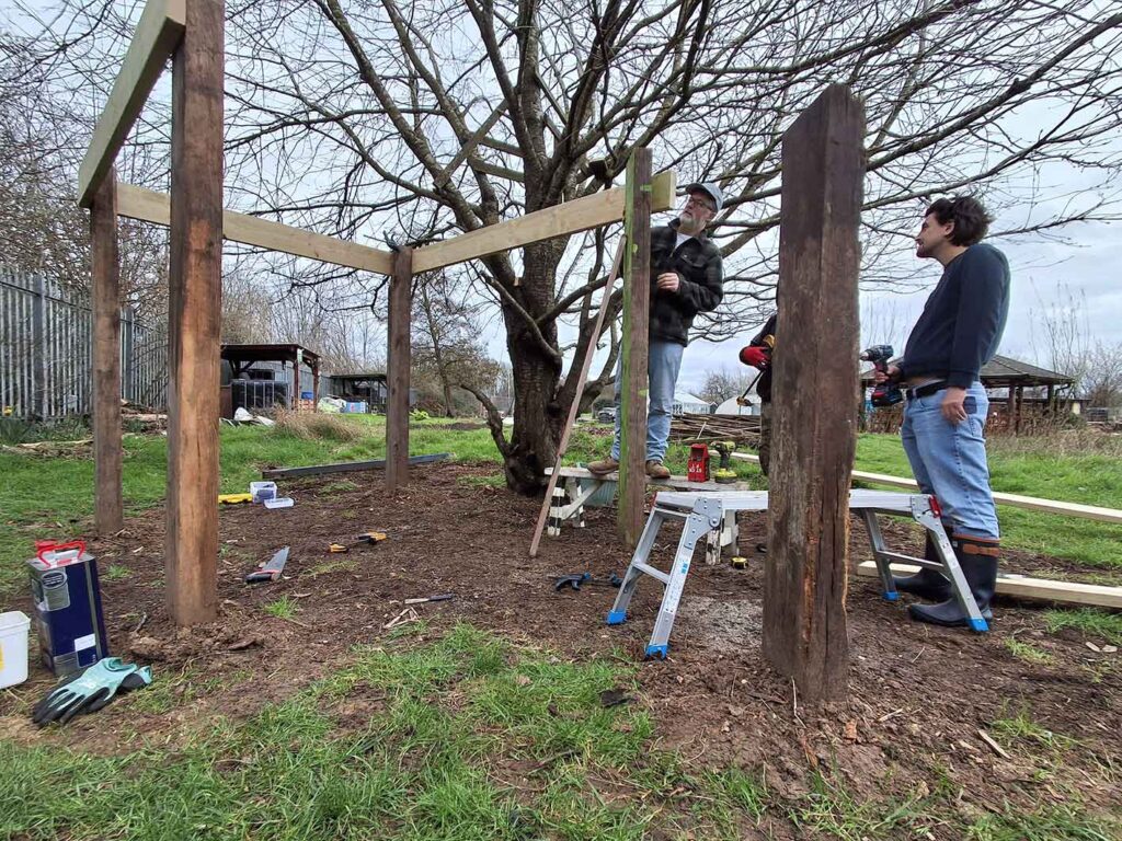 Three men working together on a wooden structure in an outdoor space. There are tools and a ladder being used in the construction.