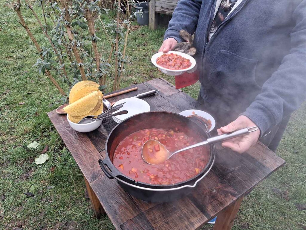 A pot of food being cooked outdoors being served up with a metal ladle into a bowl. There are taco shells on the table and steam coming off the food.