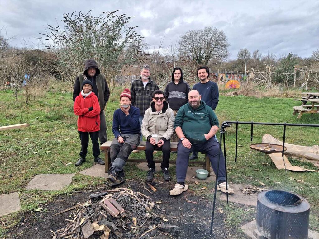 A group of men of varying ages sat and stood together around a bench in an outdoor settling with a firepit in frame. They are dressed for cold weather and are engaged with the camera.