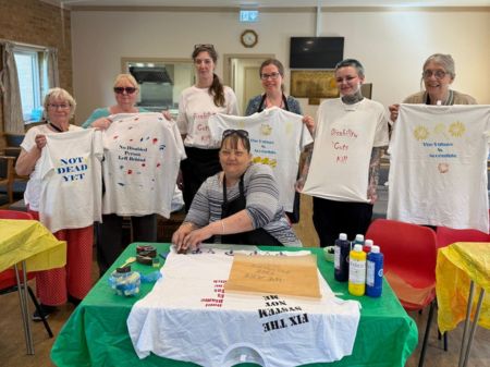 A group photo of Peterborough Sight Club wearing and showing off their protest t-shirt prints that they've made. In the centre is a green table full of workshop materials for t-shirt printing.