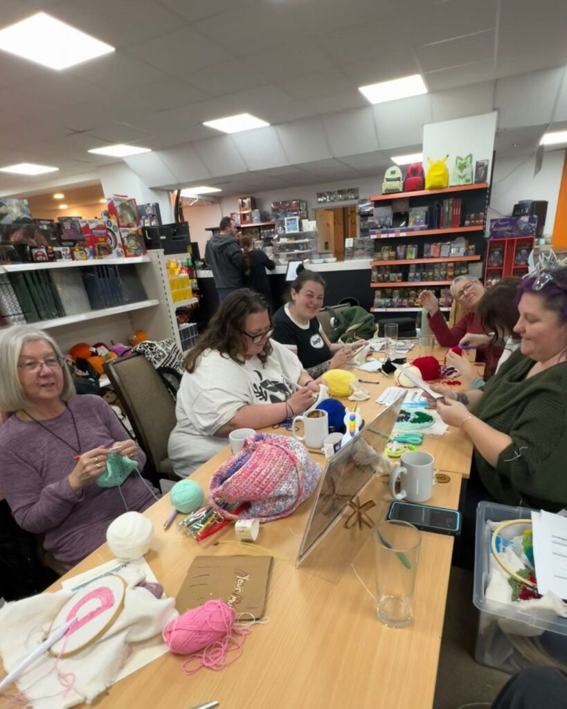 A group of people sat around a table crafting together. They are taking part in needle punch craft and there are yarn and other supplies scattered around the table. They are inside a gaming lounge and games can be seen on shelves in the background. 