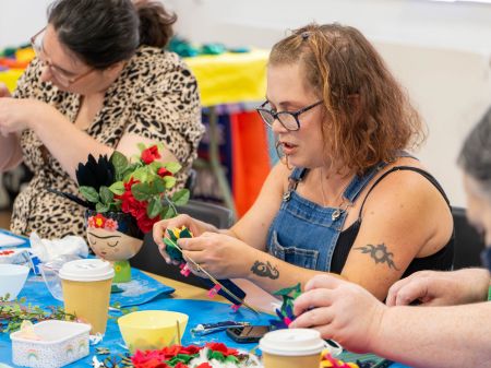 A workshop participant making their felt flower for their crown. The person has shoulder length wavy browny-orange hair, black rimmed glasses, and a tattoo on their bicep
