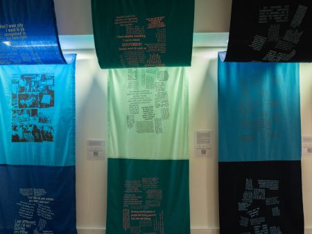 Three large fabric panels created by Fae Kilburn. The panels hang from the ceiling, draping down along the walls. The end panels are a mixture of dark and bright blues, and the middle panel is a mint green and teal. Each fabric panel is covered with black printed text.