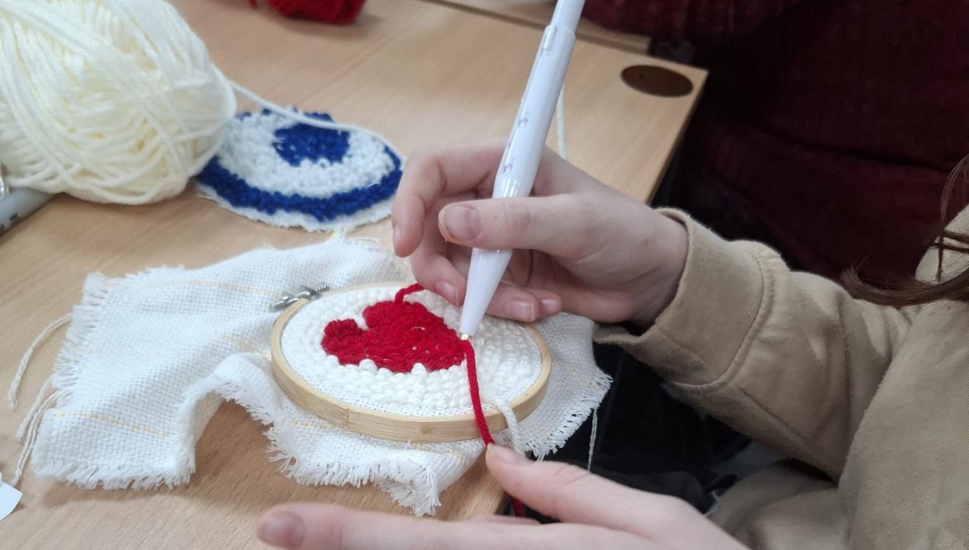 Close up of a pair of hands working on a punch needle craft design. One hand is holding the needle, working on a red heart shaped design and the other is holding the wooden craft hoop it's being stitched into. Other craft supplies are scattered in the background.