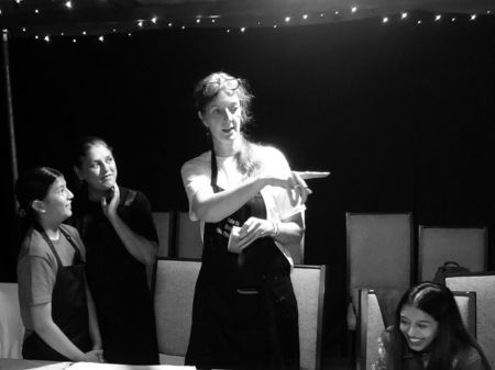 A black and white photo of Fae Kilburn delivering a workshop. Fae is stood in the middle of a family 3, pointing off-frame, as if giving instructions. The family are looking at Fae and smiling.