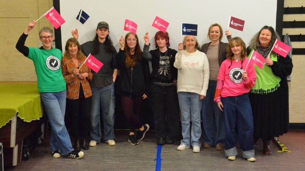 A group including young people from Eye Gets Green, staff and volunteers stood against a white projector screen holding up bright pink flags with the funder logo on. Photo by Dale McKean