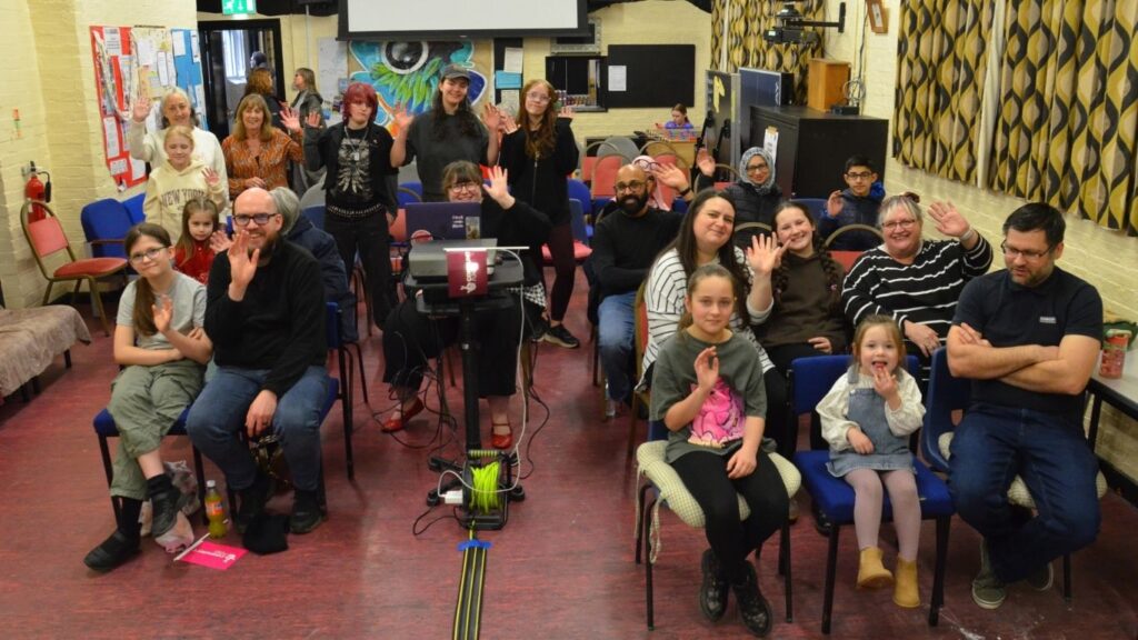A community hall full of people sat on chairs facing the front, or stood behind a projector during a film showing. Lots of young people and parents are smiling at the camera. Photo by Dale McKean