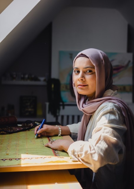 Rashida sits at a table, looking at us with a soft smile. Her hands rest of the wooden table in front of her. She wears a light dusky purple hijab and a long-sleeved cream top.