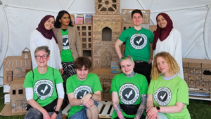 The Peterborough Presents team and volunteers wear green T-shirts with the PP logo on it. They stand in front of a cardboard city at Peterborough celebrates Festival