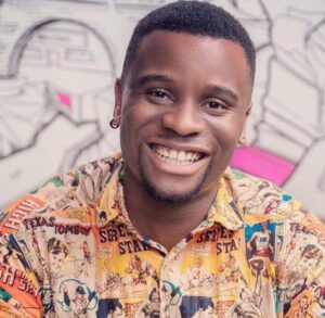 A head and shoulders image of Sonny Nwachukwu wearing a brightly patterned yellow collared shirt against a doodled background, is smiling a broad friendly smile with eye contact at the camera.