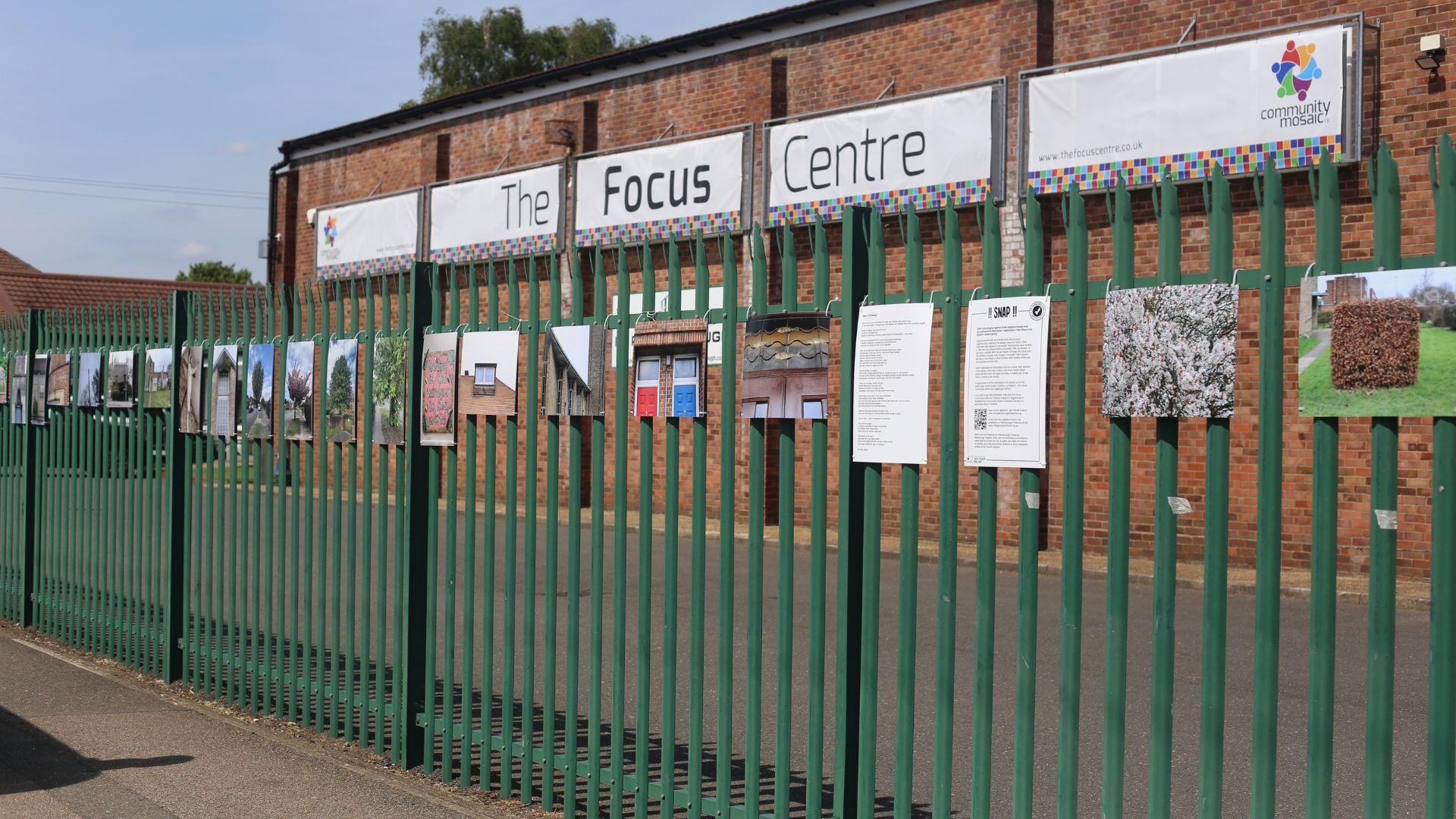 Part of the Photo Exhibition in Dogsthorpe. Large photos attached to a dark green high metal fence, with The focus Centre building in the background.