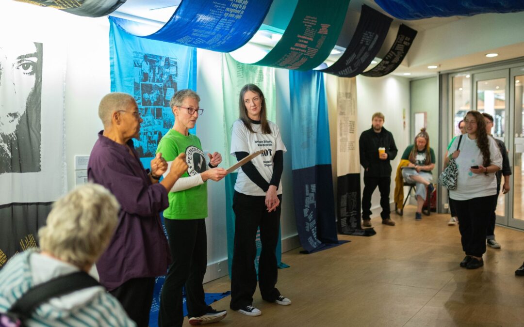 A group of people stood in an exhibition space. The three people on the left are giving a speech about the artworks in the exhibition. Kirsty (far left) is BSL intepreting, Sarah (middle, green t-shirt) is giving a speech, and Fae (right, white t-shirt with black sleeves) looks at Sarah as she is speaking. Behind them, the artworks drape from the ceiling to the floor; large fabric artworks in blues.